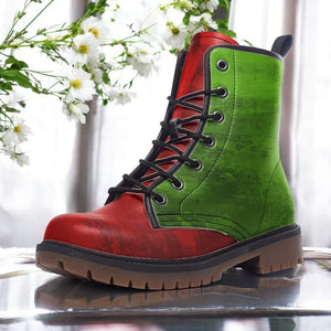 A red and green combat boot with a black lace-up design, placed on a reflective surface against a backdrop of white flowers.