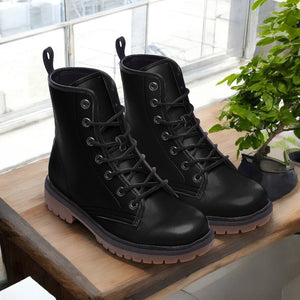 A pair of black leather boots with laces and a thick sole, placed on a wooden surface next to a potted plant.