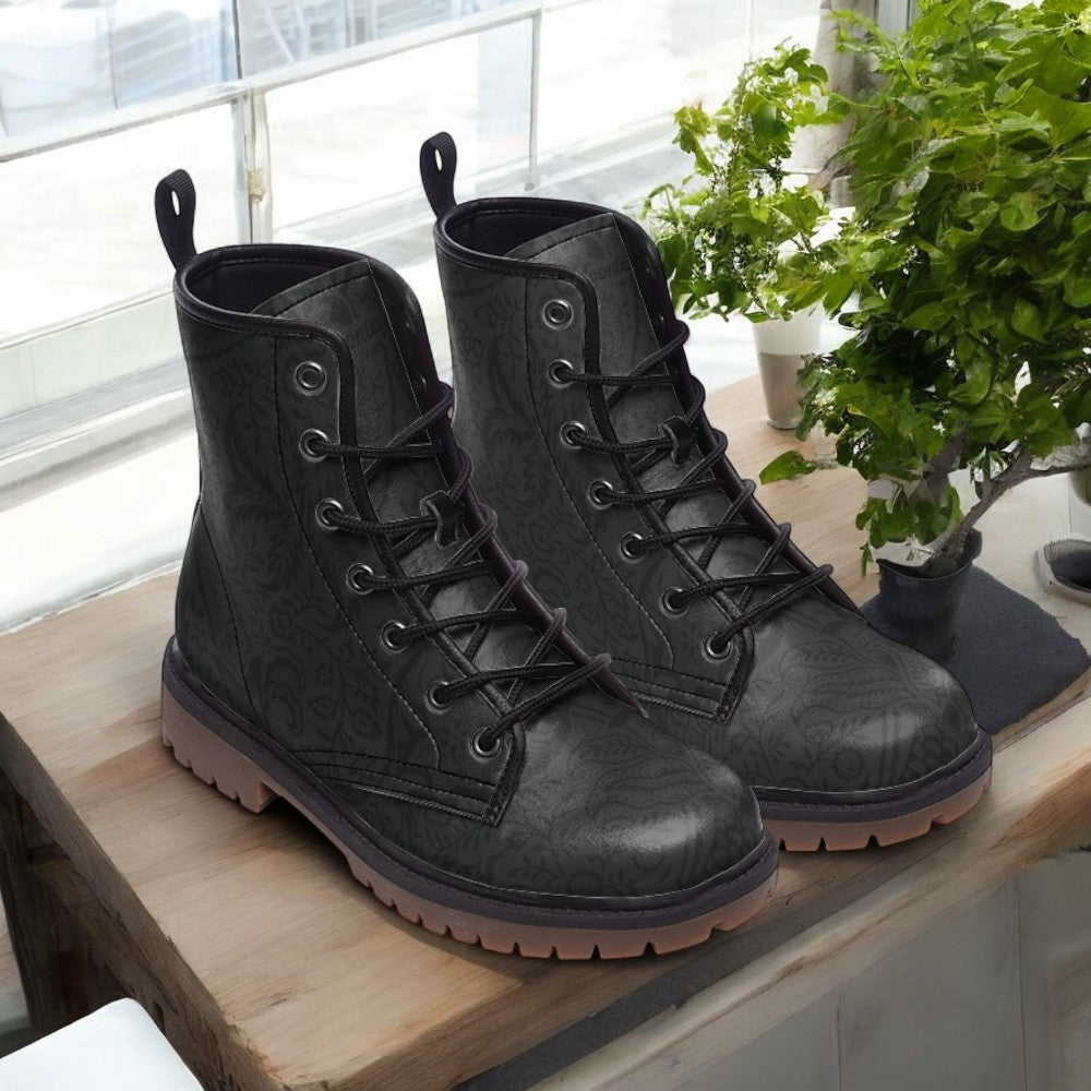 A pair of black patterned boots sits on a wooden surface, with a potted plant and a window in the background.