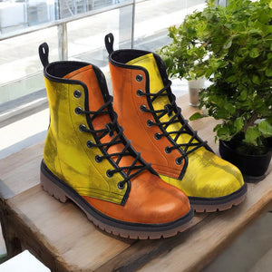 A pair of vibrant yellow and orange combat boots are displayed on a wooden surface, with a potted plant visible in the background.