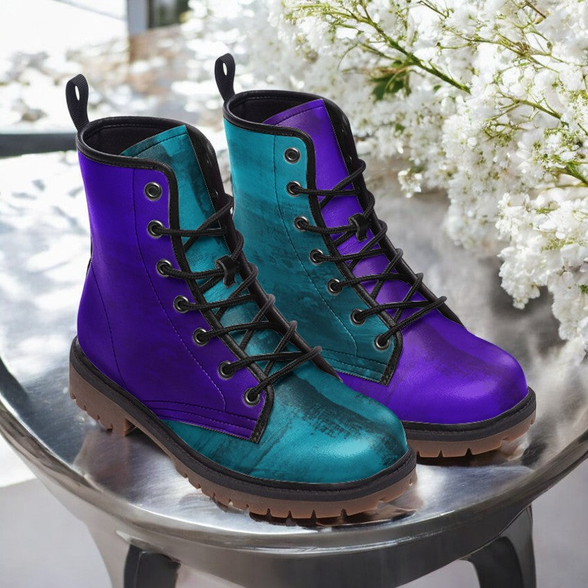 A pair of purple and teal combat boots with black laces, placed on a silver surface with white flowers in the background.