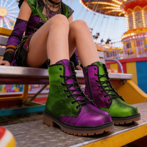 A person dressed as a clown, wearing a green and purple outfit, sits on a bench in front of a colorful carnival ride.