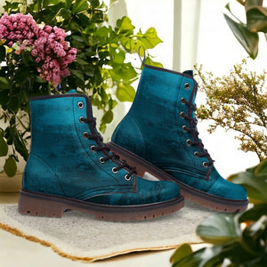 A pair of blue, textured boots with black laces, placed on a white surface with a potted plant in the background.