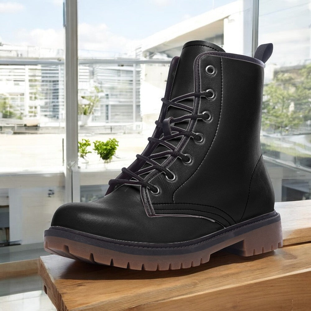 A black leather boot with a lace-up design is displayed on a wooden surface, with a window and greenery visible in the background.