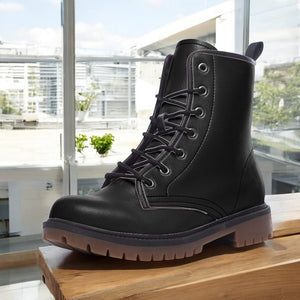 A black leather boot with a lace-up design is displayed on a wooden surface, with a window and greenery visible in the background.