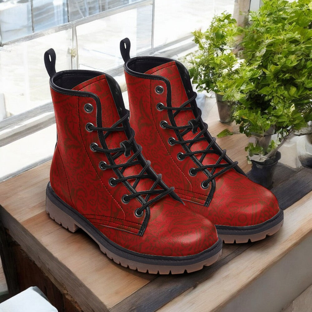A pair of red combat boots with black laces and soles, placed on a wooden surface near a window with greenery.