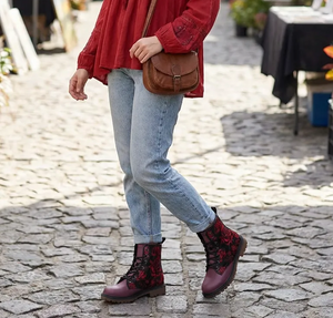 A woman wearing a red blouse, blue jeans, and red and black boots is walking on a cobblestone street, carrying a brown purse.