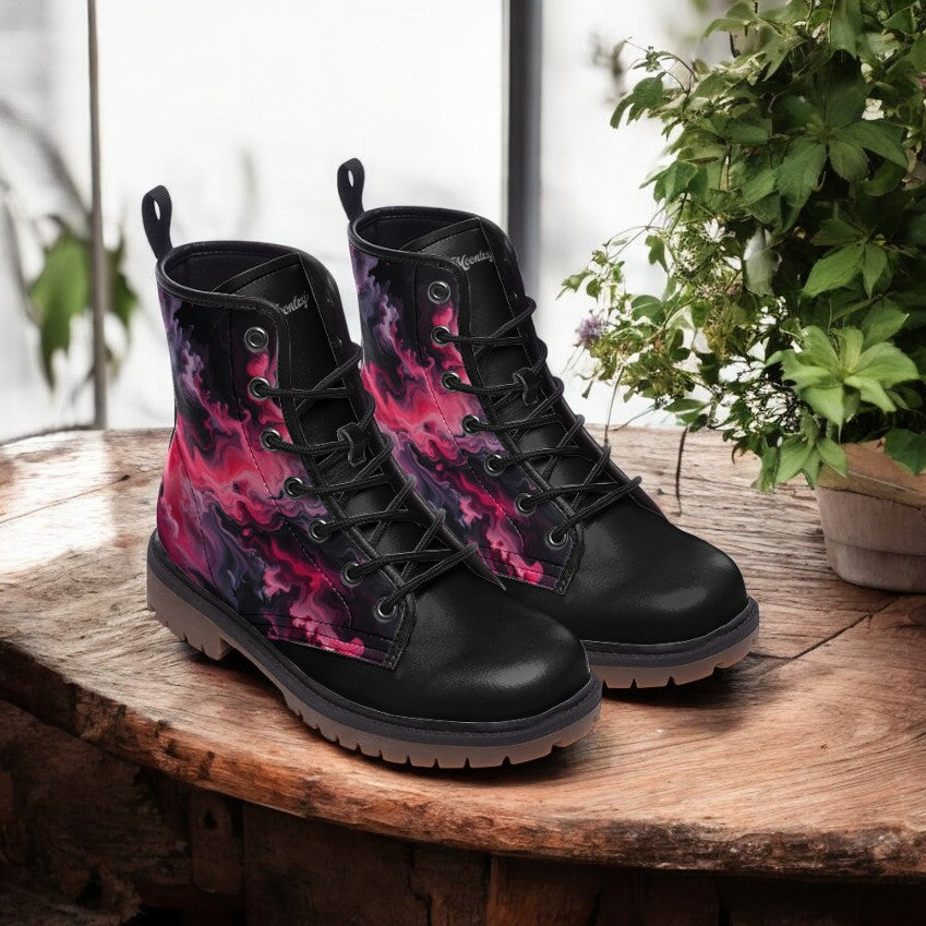 A pair of black boots with a vibrant, abstract pattern in shades of pink and purple, displayed on a wooden surface next to a potted plant.