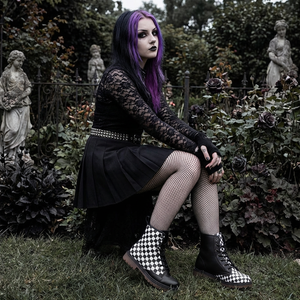 A woman with purple hair and a black lace dress sits in a garden surrounded by statues and plants.