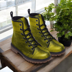 A pair of yellow combat boots with black laces and soles, placed on a wooden surface next to a potted plant.