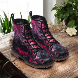 A pair of vibrant, patterned boots with a mix of pink, purple, and black colors, placed on a wooden surface near a potted plant.
