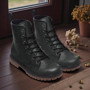 A pair of black patterned boots with laces and soles, placed on a wooden floor near a window with flowers in a pot.